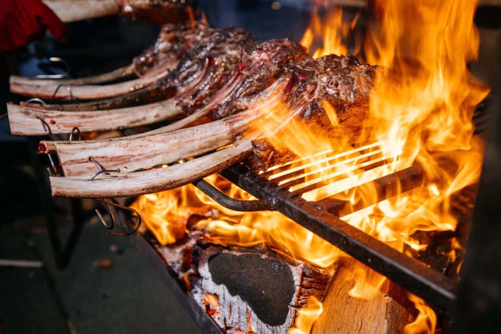 Meat on the bone sizzling on an open flame in outdoors catering event.