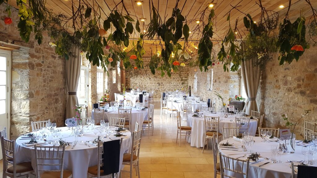a room with stone walls filled with tables with white cloths, and flowers hanging from the ceiling for a wedding catering event.