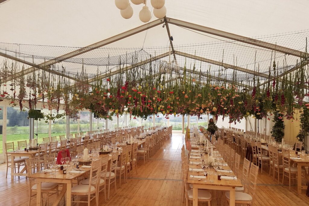 a marquee with bouquets of flowers hanging from the ceiling and tables set with glassware and serviettes underneath