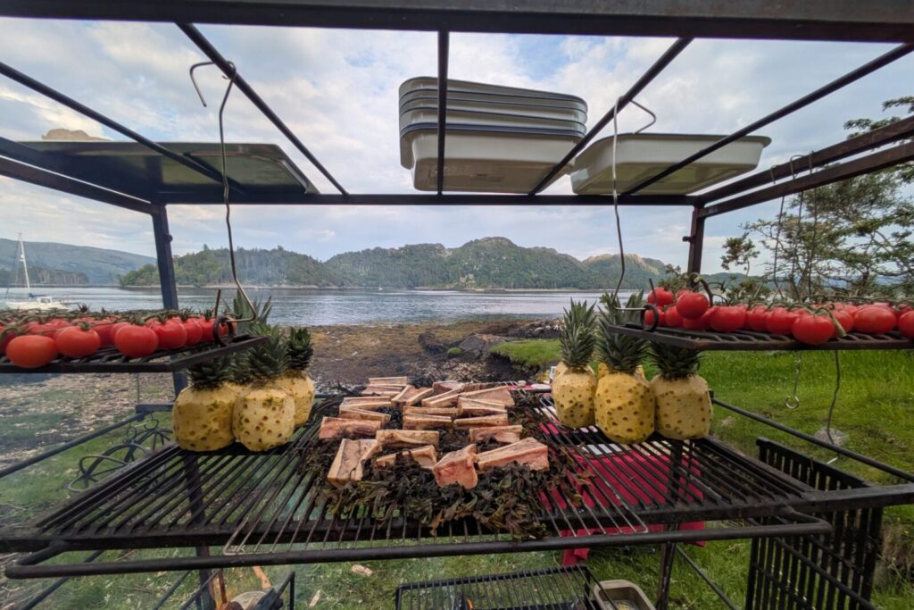 tomatoes, bones, and pineapple on a barbecue grill, which is looking out to sea at a catering event.
