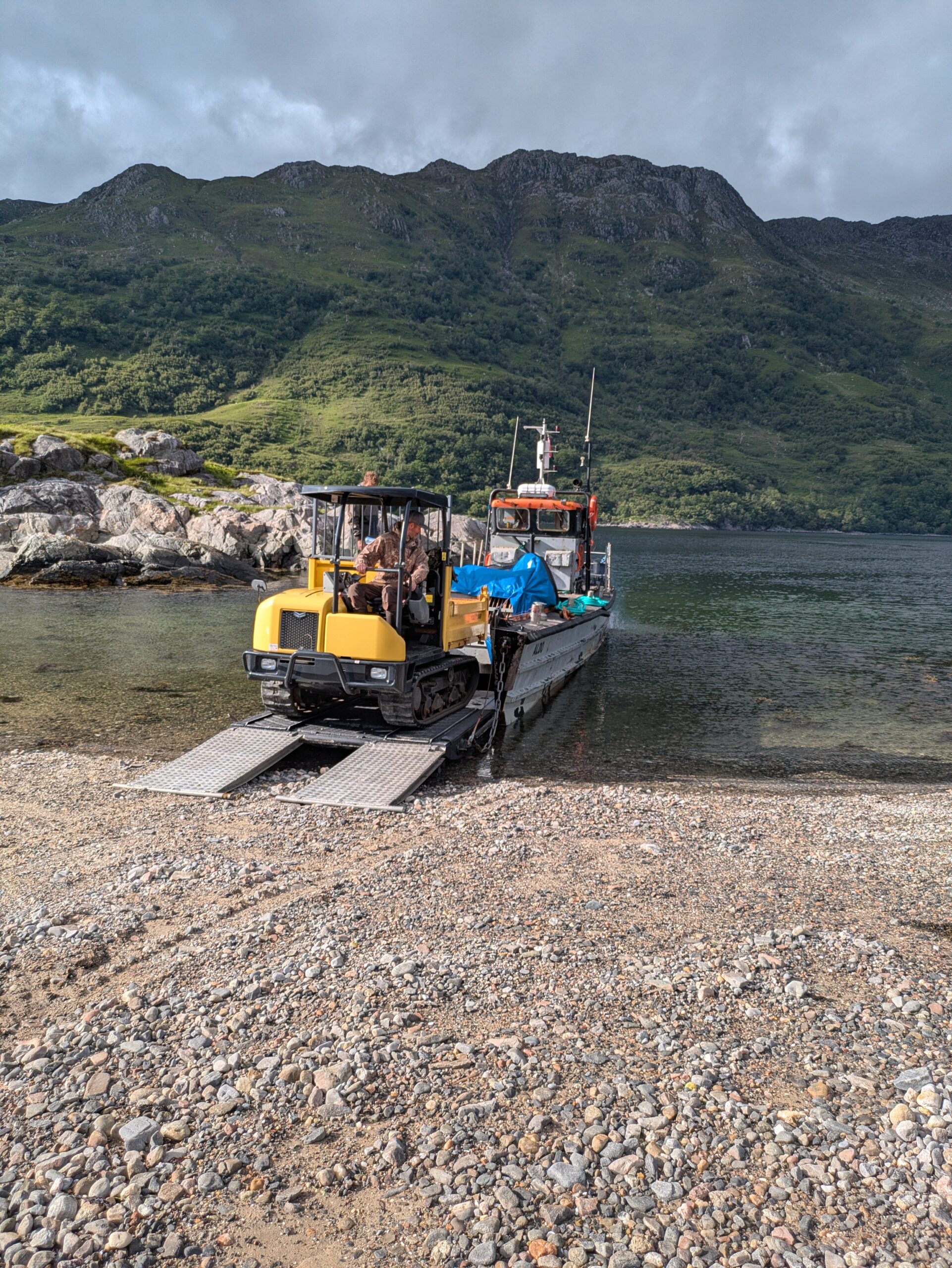 An all-terrain vehicle backs onto a small boat against rugged hills and beaches to attach a trailer with marquee event catering logistics items