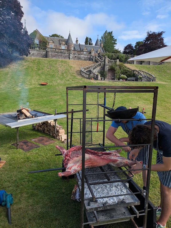 Two chefs in striped aprons prepare an asado lamb over a grill in front of a castle