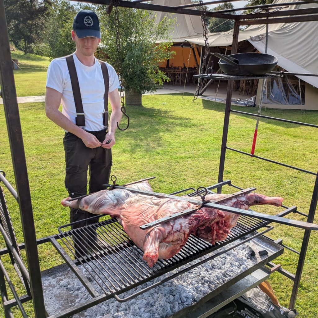 man in Ror's Kitchen cap standing in front of an asado lamb on a cross over a grill