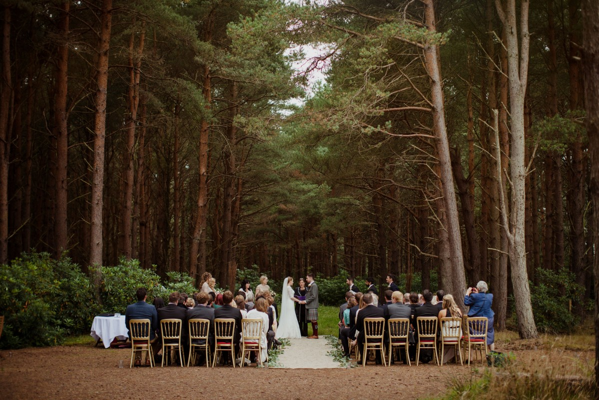 A bride and groom marrying in a forest of pines with guests watching on wooden chairs at Harvest Moon