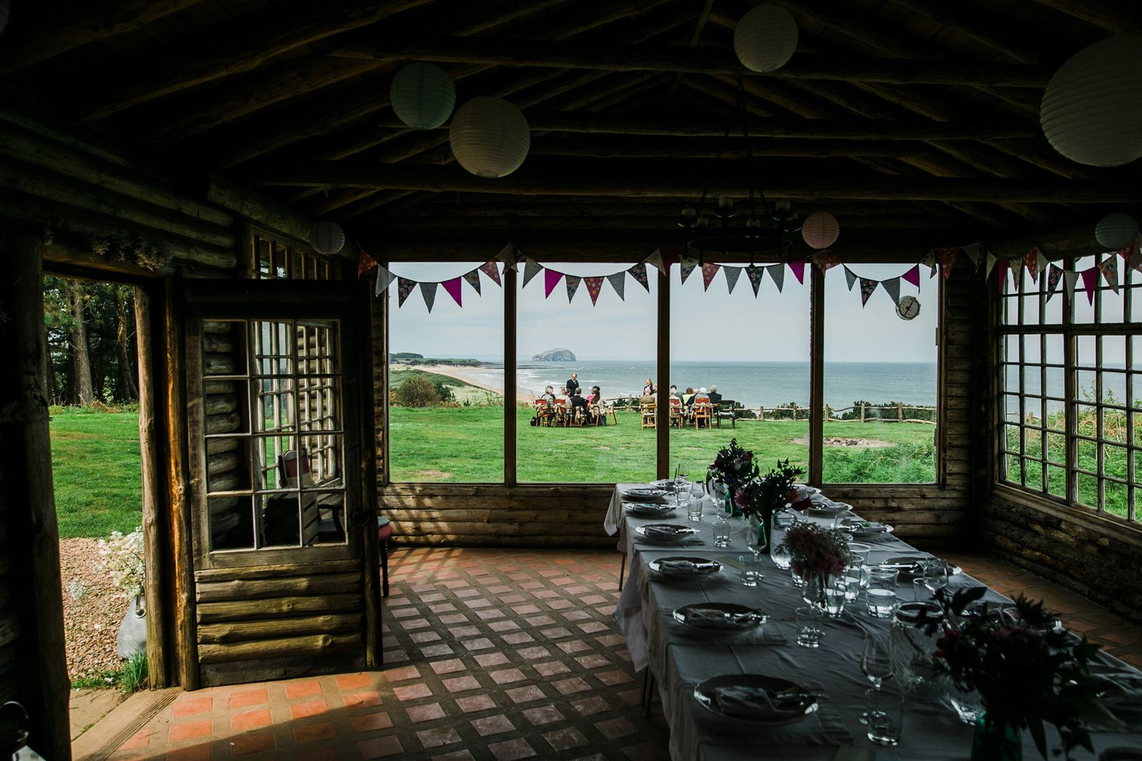 a darkened table set with plates and glasses and flowers with people sitting on chairs in front of the sea in background. Bunting.