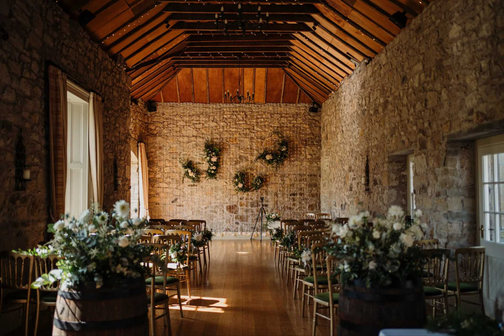 chairs line a stone room with flowers and fairy lights. The roof is wooden and it is a sunny day outside.